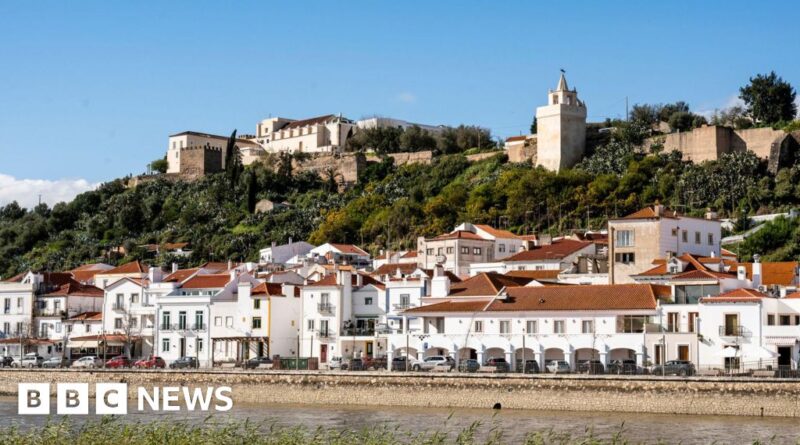 Coastal town with white houses and red-tiled roofs along the waterfront, hillside fortress in the background, BBC News watermark.
