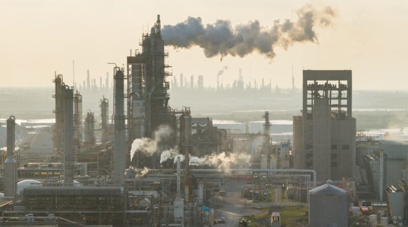 Industrial refinery complex with tall smokestacks emitting smoke against a hazy sky