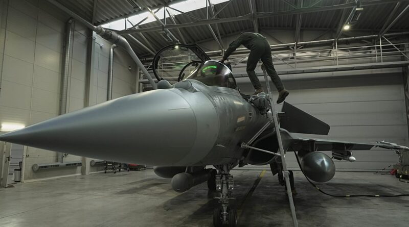Maintenance crew member climbs a ladder to access the cockpit of a gray fighter jet in a hangar