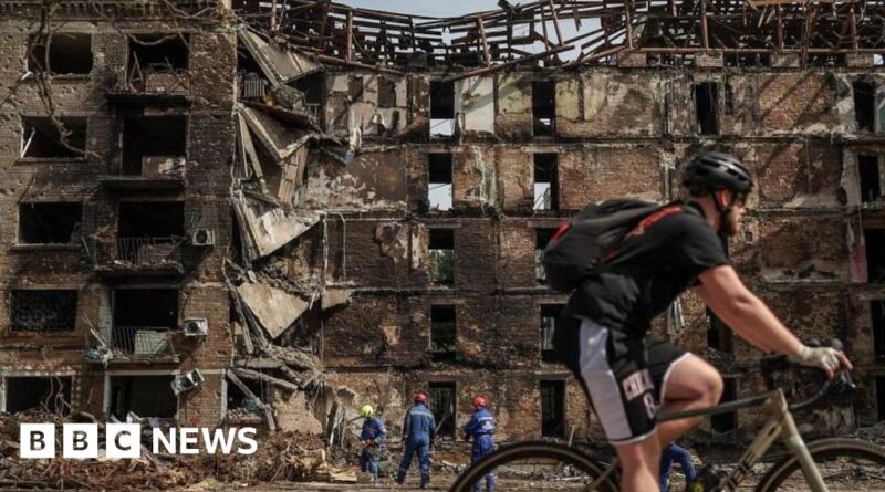 Cyclist riding past a heavily damaged brick building with collapsed upper floors as workers in helmets examine rubble in the background.