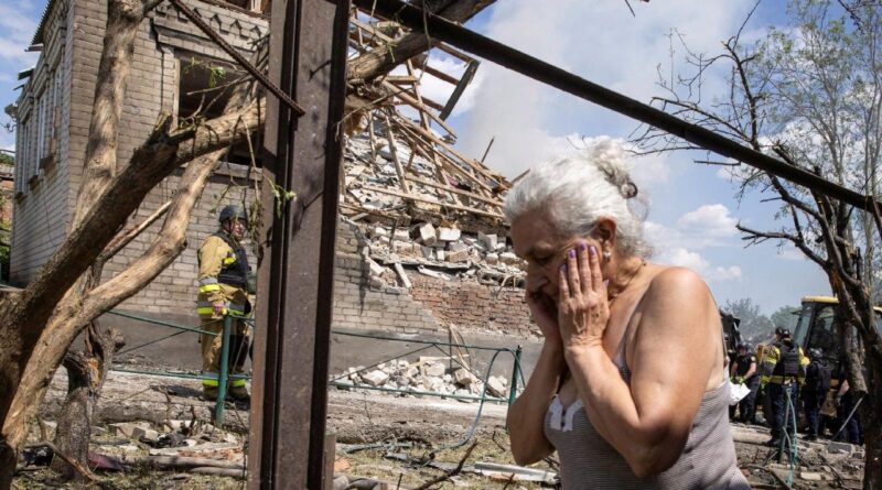 Elderly woman with gray hair trembling, hands to face, amid rubble and a partially collapsed building with firefighters in the background.