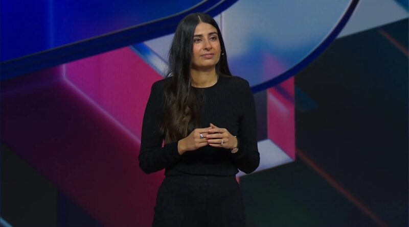 Female presenter in a black long-sleeve top stands on a colorful stage, hands clasped while speaking.