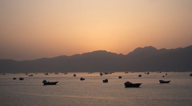 Sunset over a calm bay with silhouetted boats and distant mountain silhouettes on the horizon.