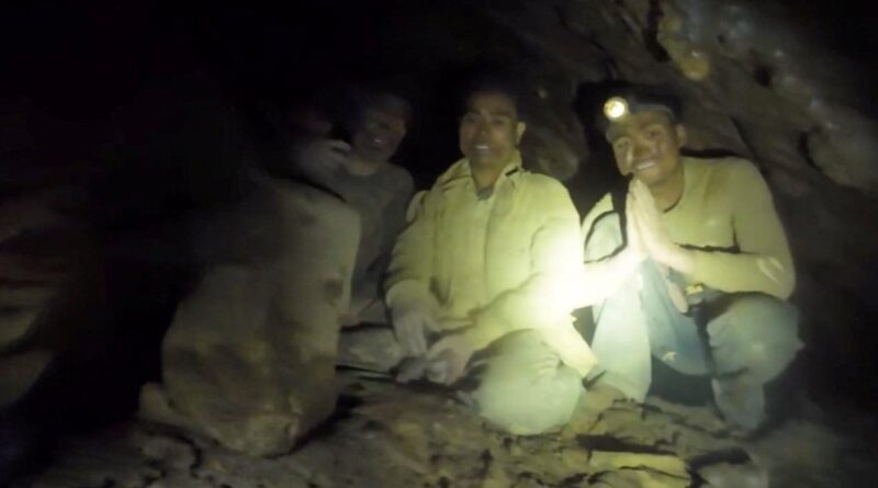Three men sit on a rocky cave floor, one wearing a headlamp, with the middle man smiling and two others nearby in dim light.