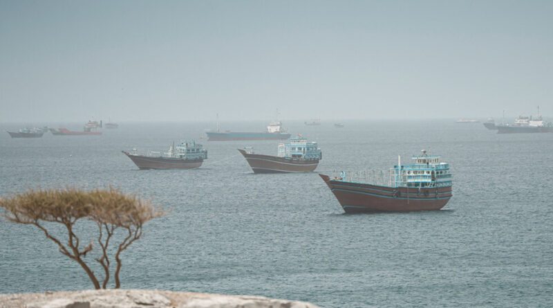 Calm sea with several blue and white fishing boats anchored across a hazy horizon, a lone tree at the left foreground.