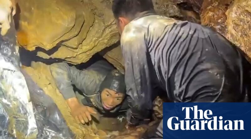 Two cavers in a narrow, muddy cave passage; one wearing a helmet, the other close behind.