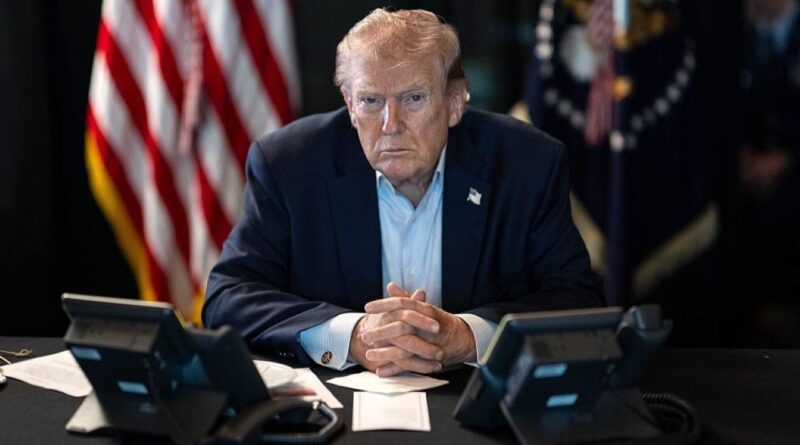 Man in a dark suit sits at a conference table with two phones and papers, hands clasped, American flag in the background.