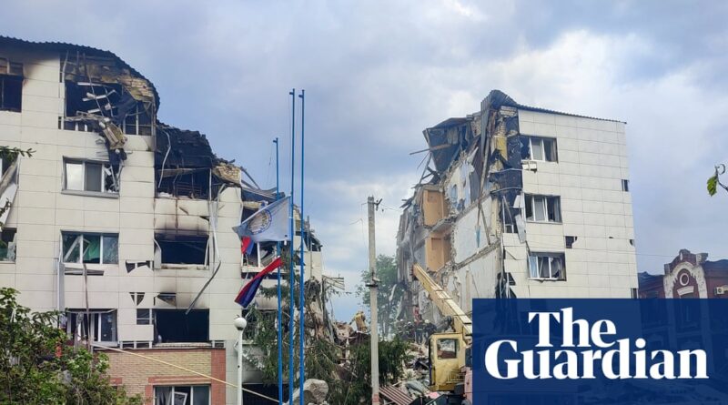 Collapsed apartment building with exposed interiors and rubble after a disaster; rescue crews and a crane amid debris under a cloudy sky.