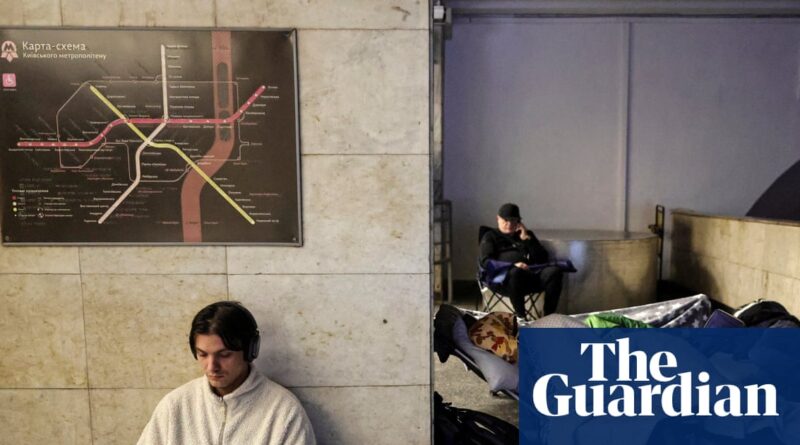 Man with headphones sits against a tiled wall beneath a subway map; a makeshift camp with blankets and a second passerby is visible nearby.