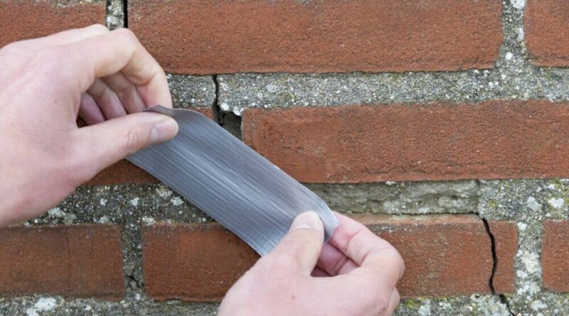 Hands press a thin gray metal repair strip onto a crack in a brick wall.