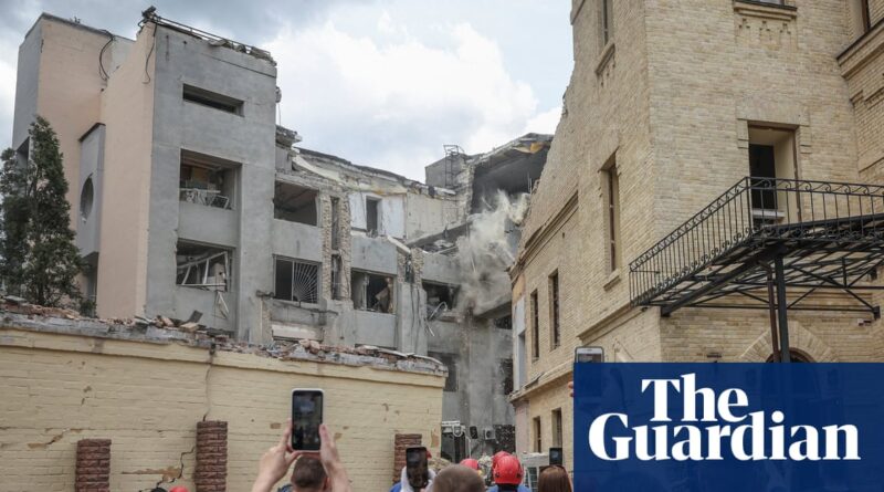 Partially collapsed multi-story building with exposed interiors and smoke as onlookers film with their phones.