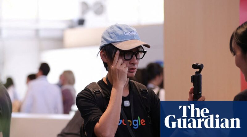 Young man wearing a light blue Google cap and glasses, touching his temple at a busy event with a Guardian watermark in the corner