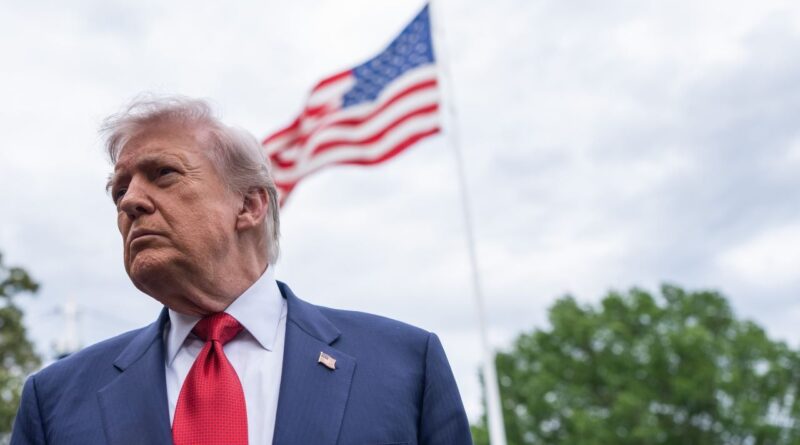 Man in a blue suit and red tie outdoors, with an American flag waving in the background.