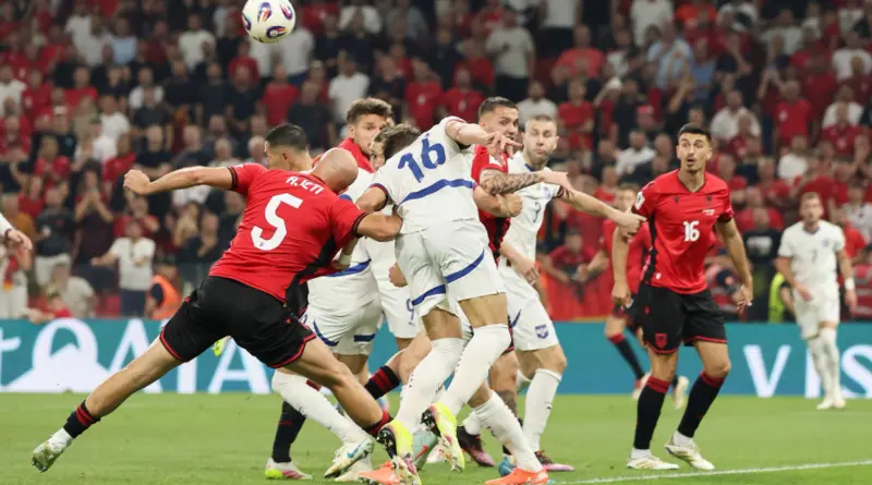 Soccer players in red and white kits scramble for the ball in front of the goal as the crowd watches intently from the stands.