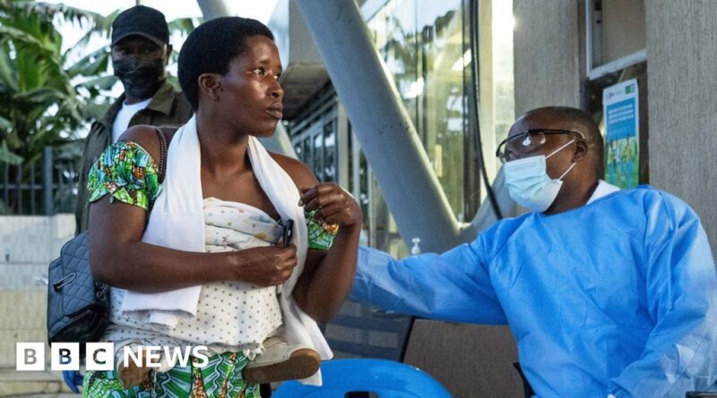 Healthcare worker in blue PPE and mask reaches toward a seated woman holding a wrapped baby outdoors by a glass building.