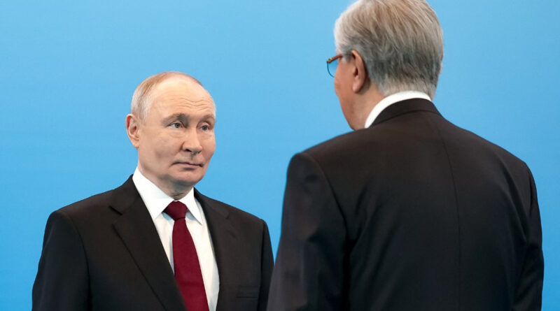 Two men in dark suits and red tie stand against a blue backdrop, facing each other in a formal setting with neutral expressions.