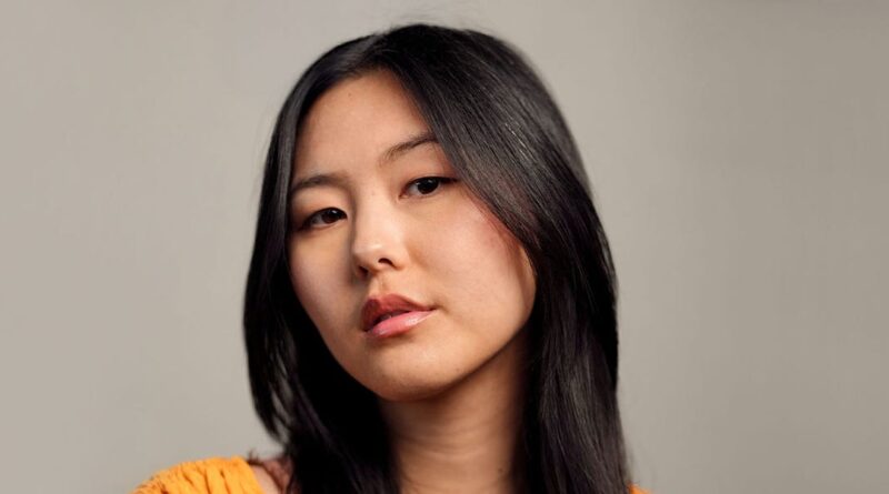 Portrait of an Asian woman with long black hair, wearing an orange top, looking softly at the camera against a neutral gray background.