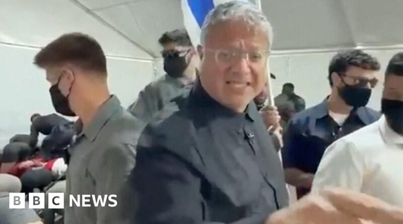 Central man with gray hair in a black shirt smiles at the camera, surrounded by masked people inside a tent.