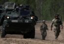Armored personnel carrier on a dusty road, flanked by two soldiers in camouflage.