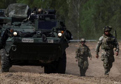 Armored personnel carrier on a dusty road, flanked by two soldiers in camouflage.