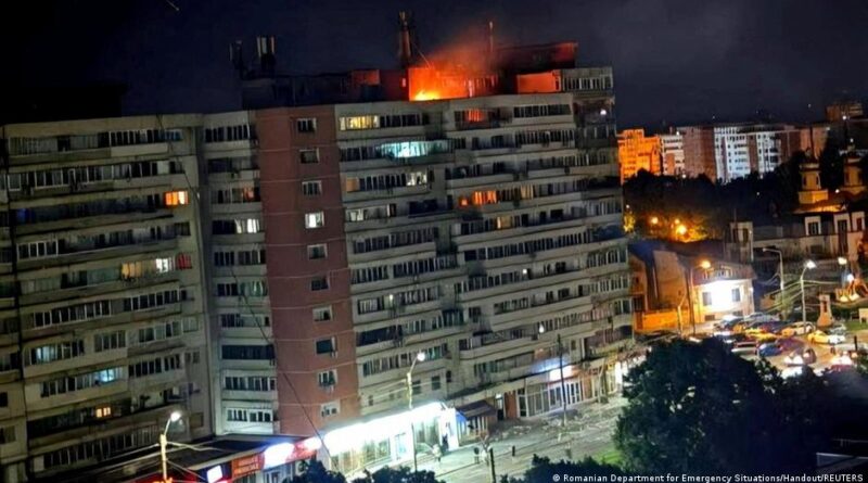 Night view of a multi-story apartment building with a large fire burning on the roof while city lights glow below.