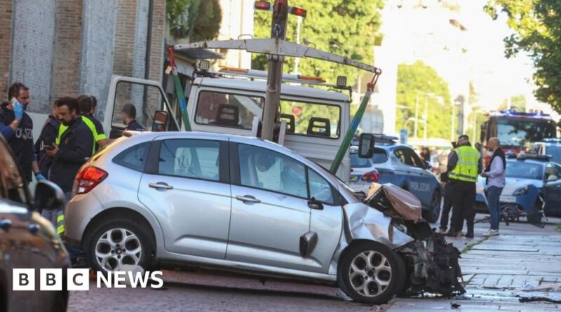 Silver hatchback severely damaged in a crash on a city street; tow truck arm above the car as police and responders assist nearby.