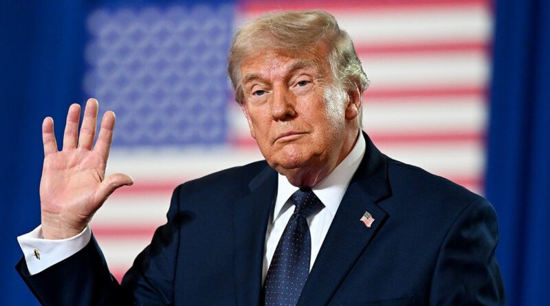 Older man in a dark suit waves with his right hand, with a blurred American flag behind him.