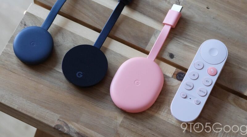 Three Google Chromecast dongles in blue, black, and pink with a white remote on a wooden table.