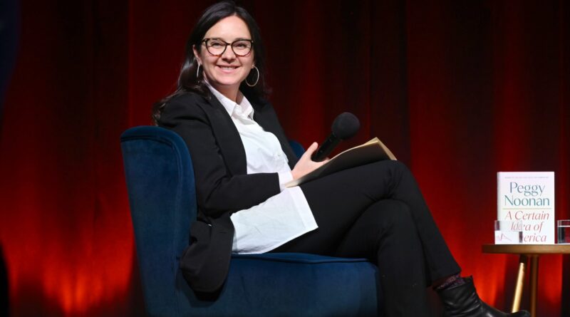 Speaker in a black blazer with white shirt sits in a blue chair on stage, holding a microphone and a notebook, with a book on a small table beside her.