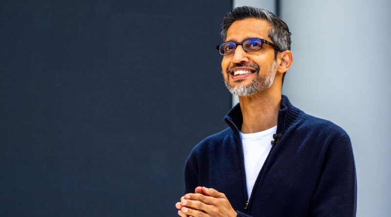 Portrait of a smiling man with glasses and gray beard, wearing a navy zip-up sweater and a lapel mic, standing against a dark backdrop up to the right side of the frame.