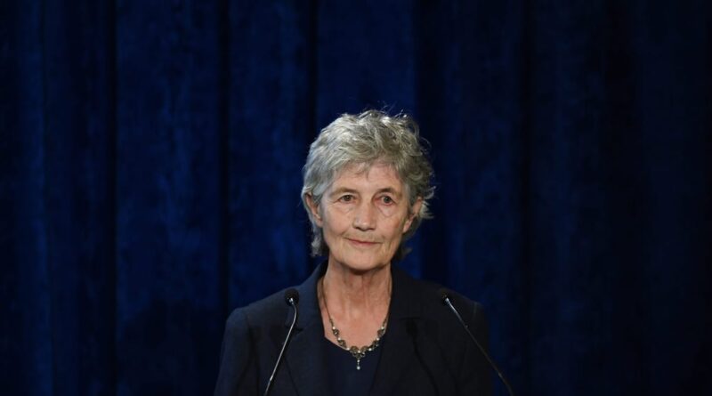 Older woman with short gray hair speaking at a podium against a dark blue curtain backdrop.