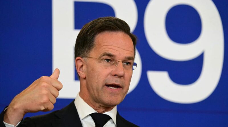 Man in a dark suit and glasses giving a thumbs-up during a speech, with a blue background and large white numbers behind him.