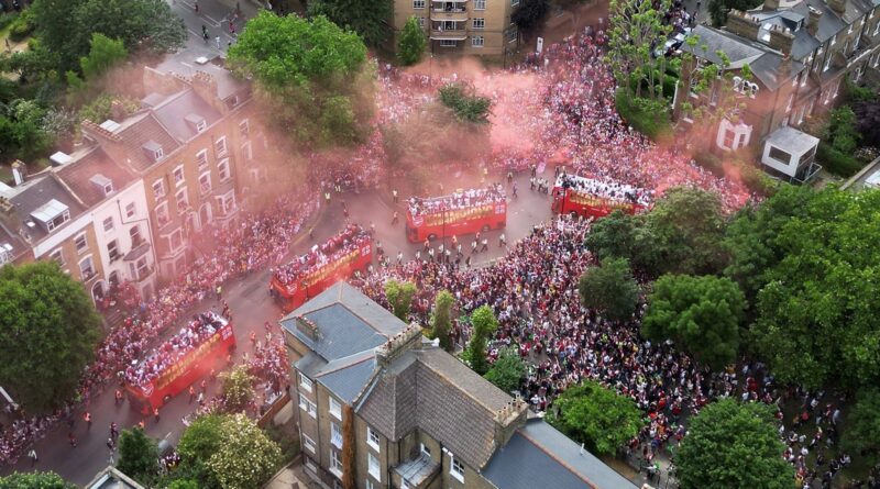 Aerial view of a crowded city street parade with red buses, pink smoke, and dense cheering crowds among trees and buildings.