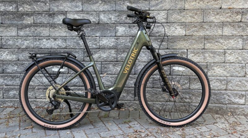 Olive-green Velotric e-bike parked against a stone wall with tan tires and a rear rack on the back wheel side of the frame.