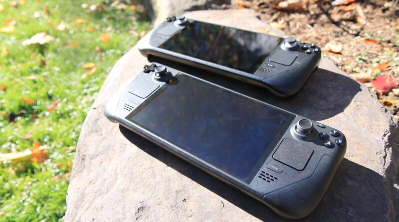 Two handheld gaming consoles resting on a rock outdoors with grass and fallen leaves nearby, sunlight on the devices.
