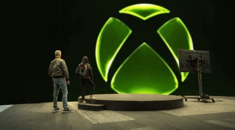Two people stand on a circular stage, facing a massive neon green Xbox logo projected on a dark wall with a monitor on a stand to the right.