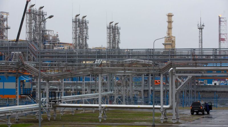 Extensive network of metal pipes and industrial towers at an oil refinery, with a dark SUV parked on a wet, paved area in the foreground.