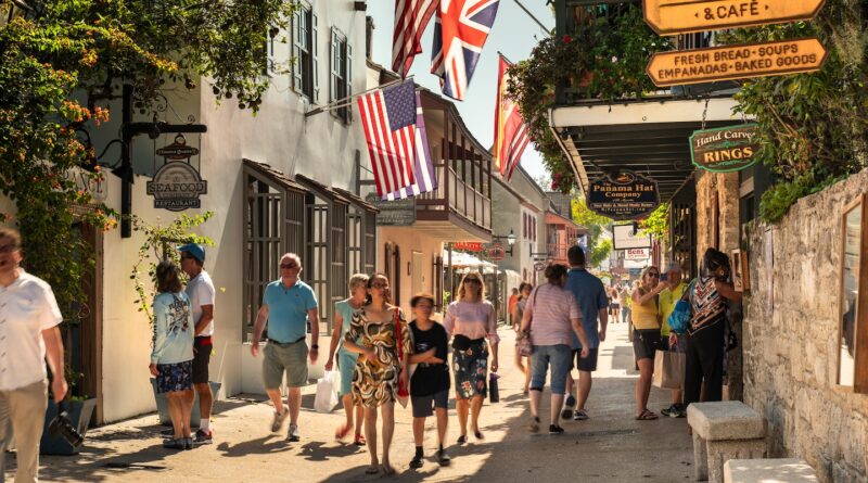Busy pedestrian street with flags overhead, storefronts on both sides, and people shopping in a sunny historic town.