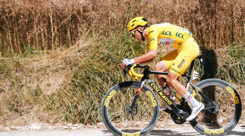 Cyclist in a bright yellow racing kit and helmet riding a carbon road bike along a dusty rural road with dry grass in the background (race number 11 visible).