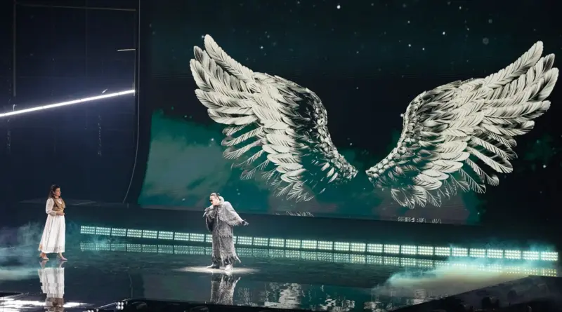 Two performers on a glossy stage with a large screen showing white feathered angel wings behind them.