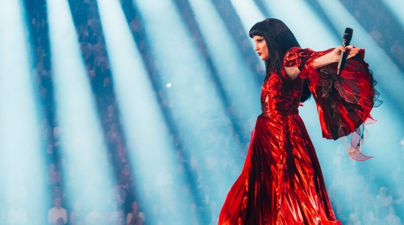 Female singer in a shiny red gown performing on stage, holding a microphone under blue spotlight beams
