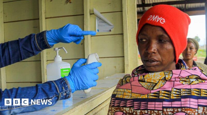 Woman in a colorful wrap and red beanie receives hand sanitizer from gloved hands at a counter, BBC News watermark visible.
