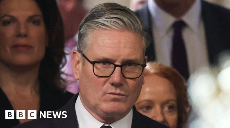 Man wearing glasses in a suit at a BBC News event, looking serious with a crowd in the background.