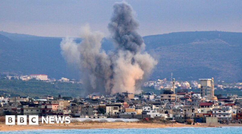 Large gray smoke plume rising from a blast in a coastal city, with buildings and hills in the background.