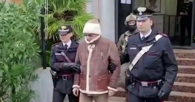 Man in a brown jacket and white beanie being escorted by two police officers down the steps outside a building.