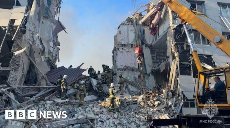 Firefighters and emergency workers search rubble at a collapsed building site with a yellow crane nearby.
