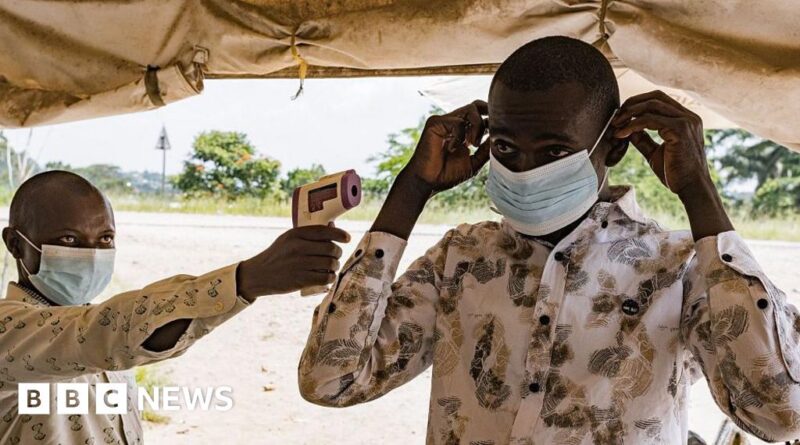Health worker in a mask uses a handheld infrared thermometer to check a man's temperature under a tent, BBC News logo visible.