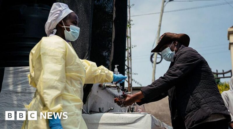 Healthcare worker in PPE and mask assists a man washing his hands at an outdoor sink.