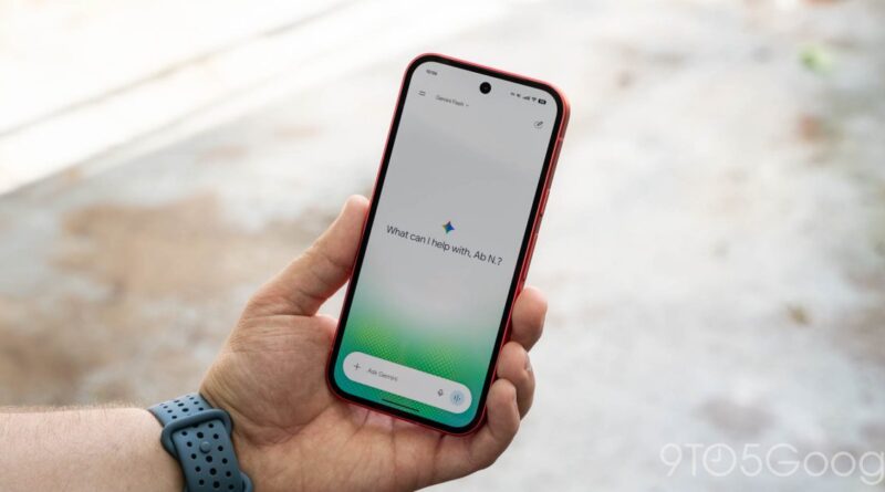 Hand holding a red-edged smartphone displaying Google Assistant home screen outdoors, with a blurred outdoor background.