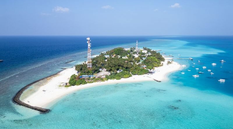 A small tropical island with white sandy beaches, dense green trees, and several antenna towers, surrounded by turquoise water and moored boats.